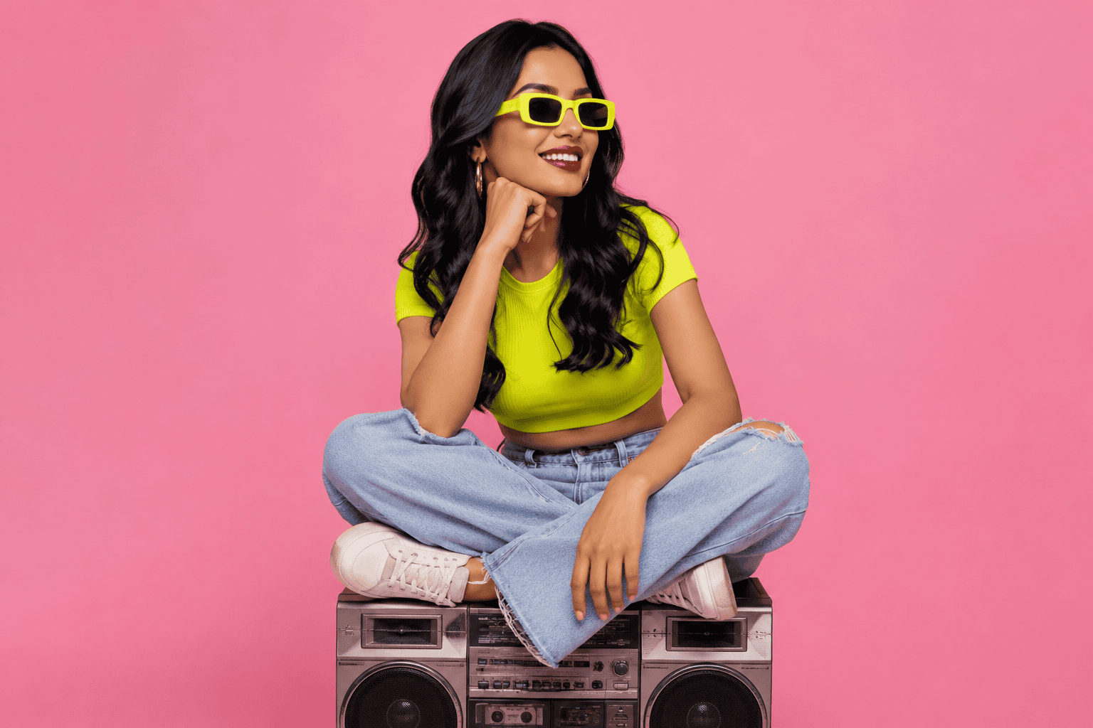 Full-body studio portrait of a smiling woman with long dark hair sitting cross-legged on a vintage silver-and-black boombox against a solid pink background