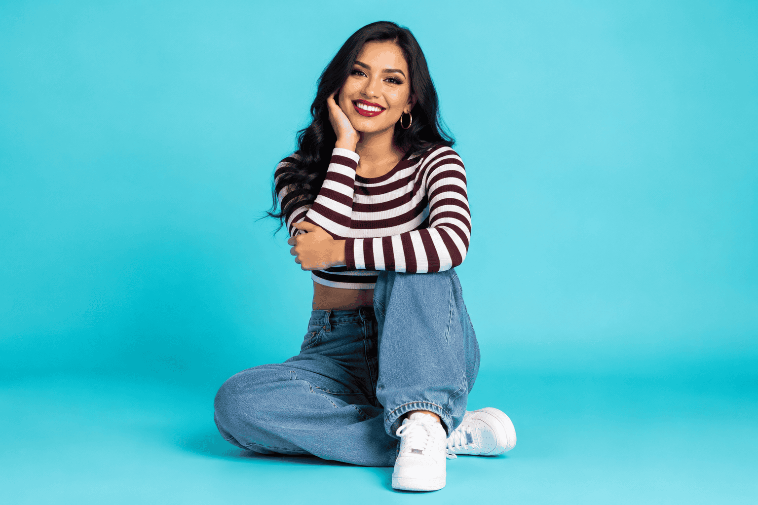 Full-body studio portrait of a smiling woman in a striped crop top, blue baggy jeans, and white sneakers, sitting against a solid turquoise background.