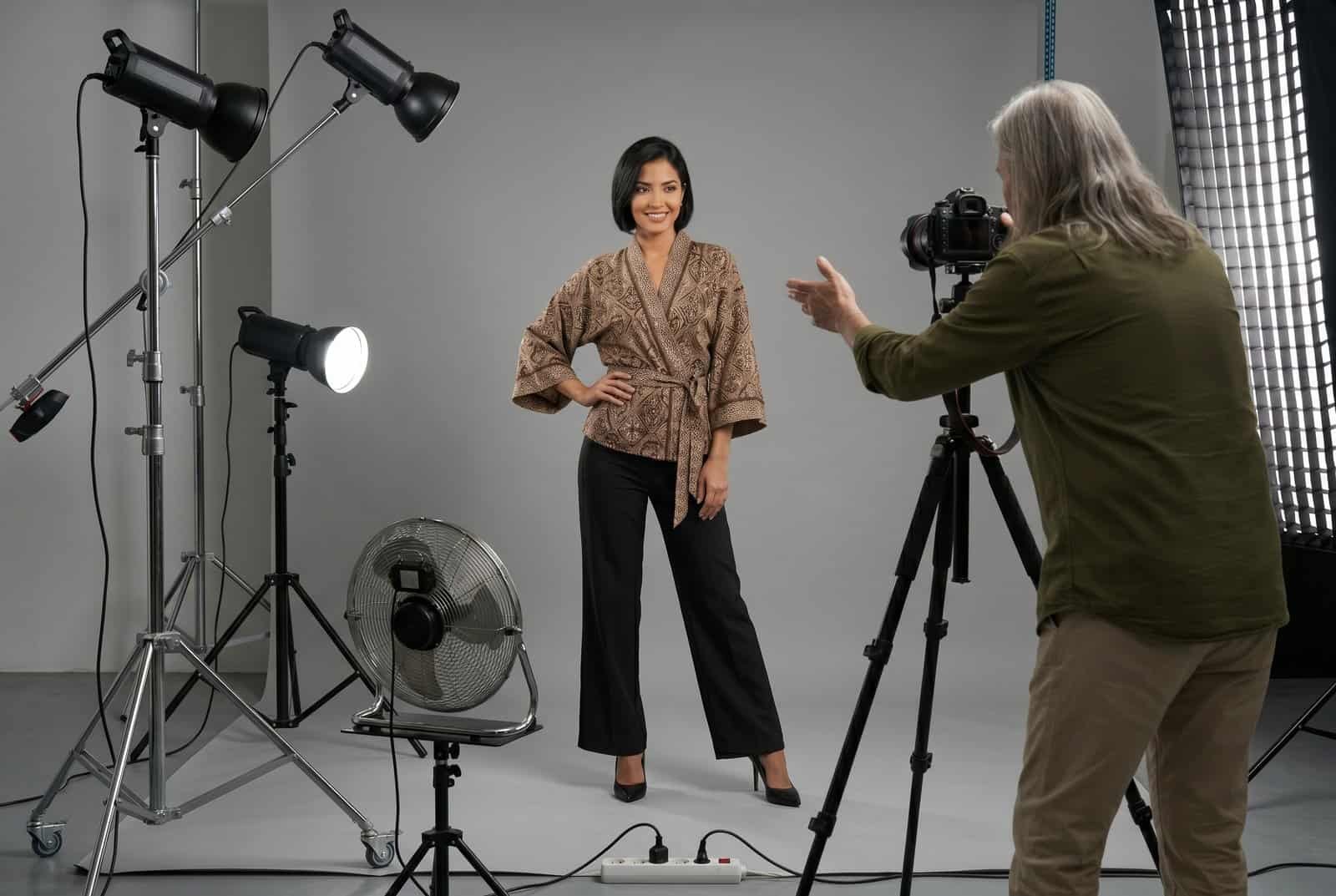 A young female model in a patterned kimono jacket posing in a professional photography studio while an older photographer directs her from behind a camera.