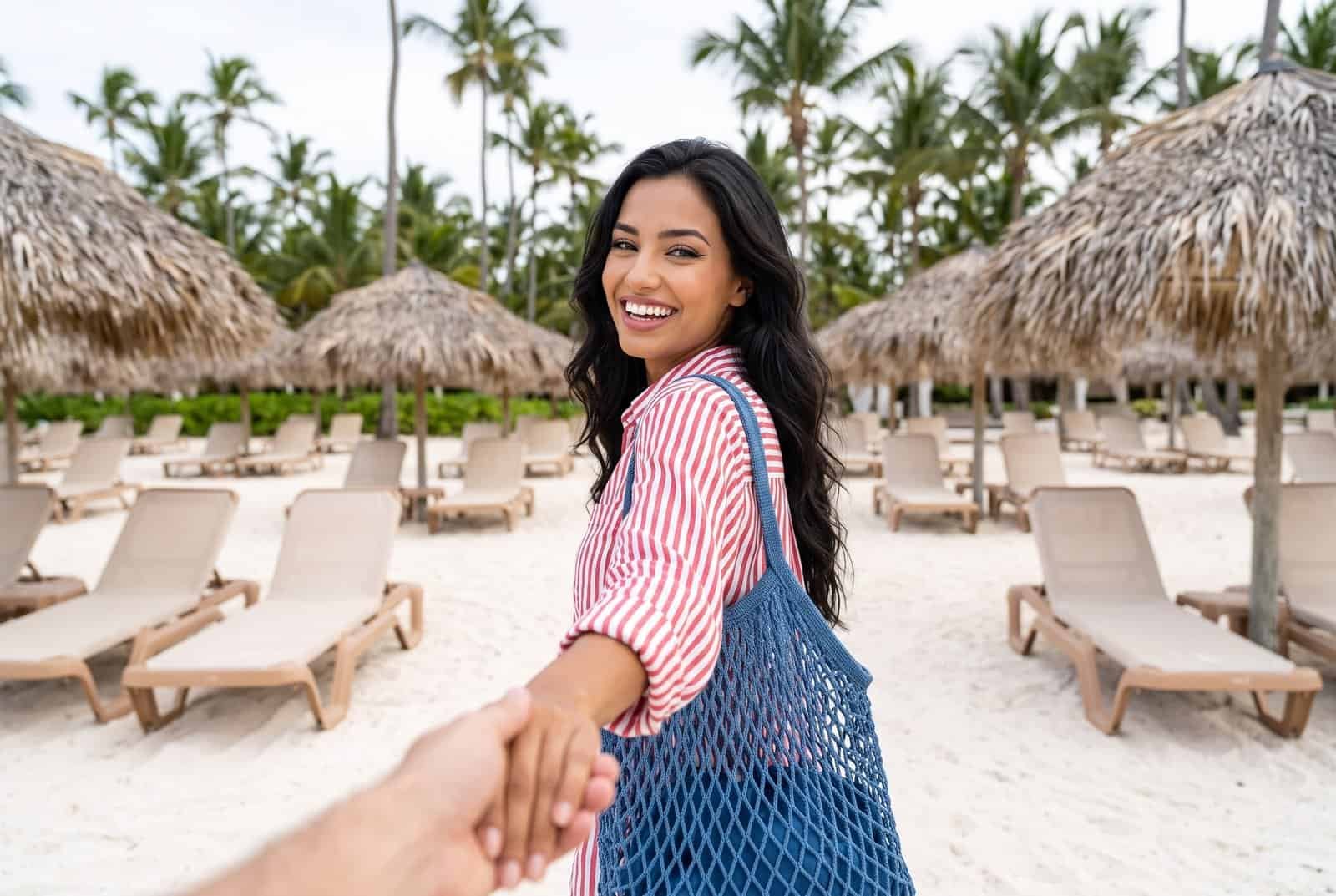 A first-person view of a smiling woman in a red and white striped shirt looking over her shoulder, holding the viewer's hand on a beautiful tropical beach with white sand, palm trees, and thatch-roof umbrellas.