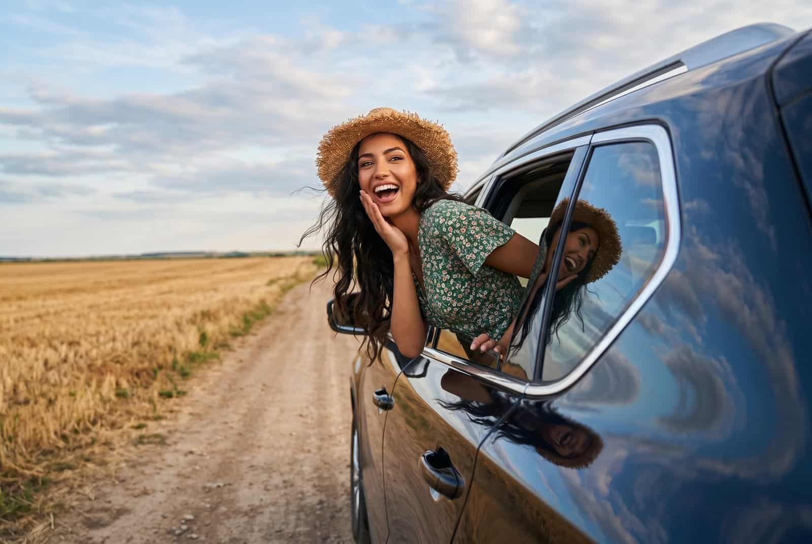 A joyful young woman in a straw hat leaning out of an SUV window next to a golden field.