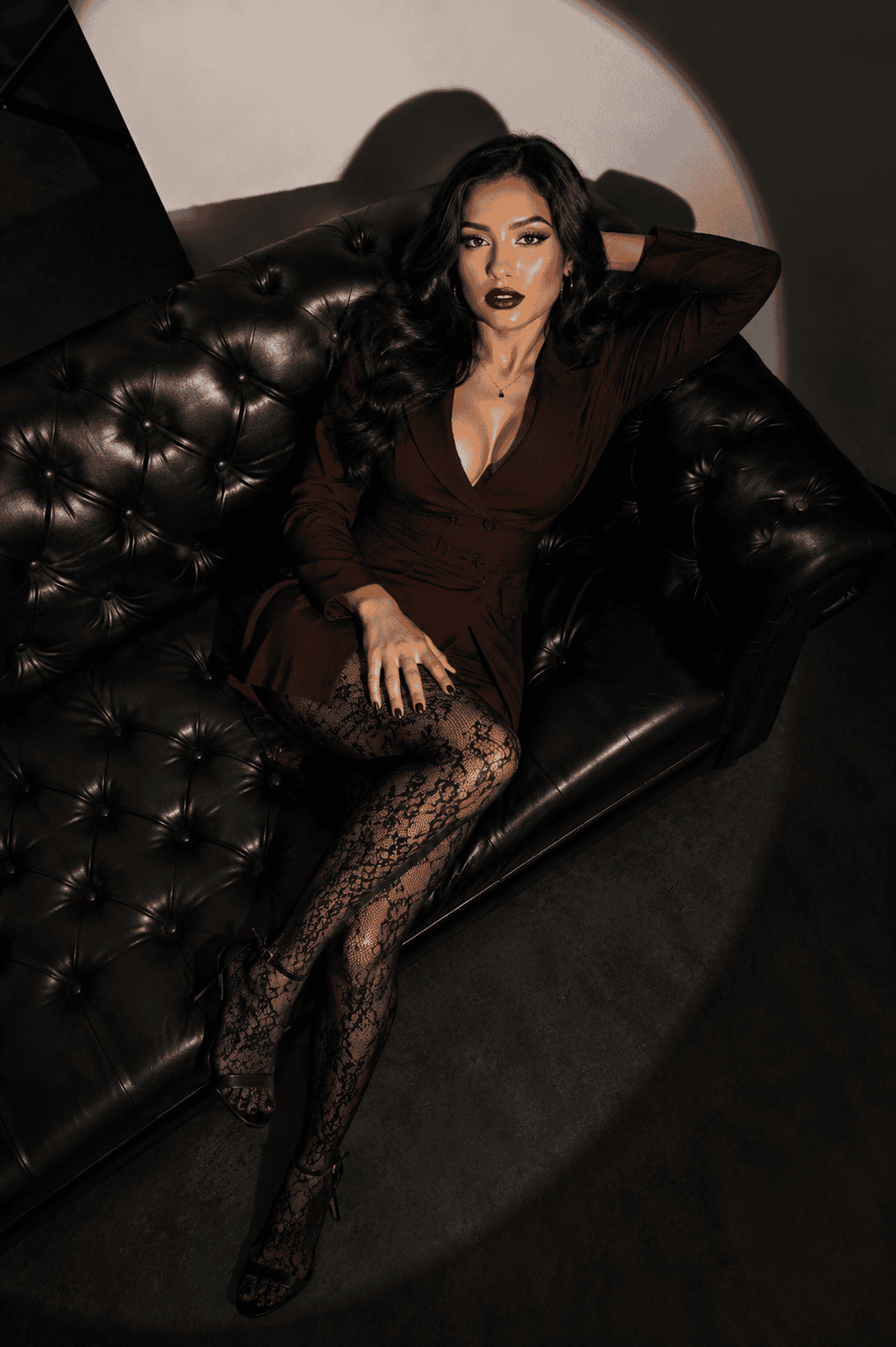 Woman reclining on a black leather sofa in a dark studio, wearing a brown blazer dress with lace tights under dramatic spotlight lighting.