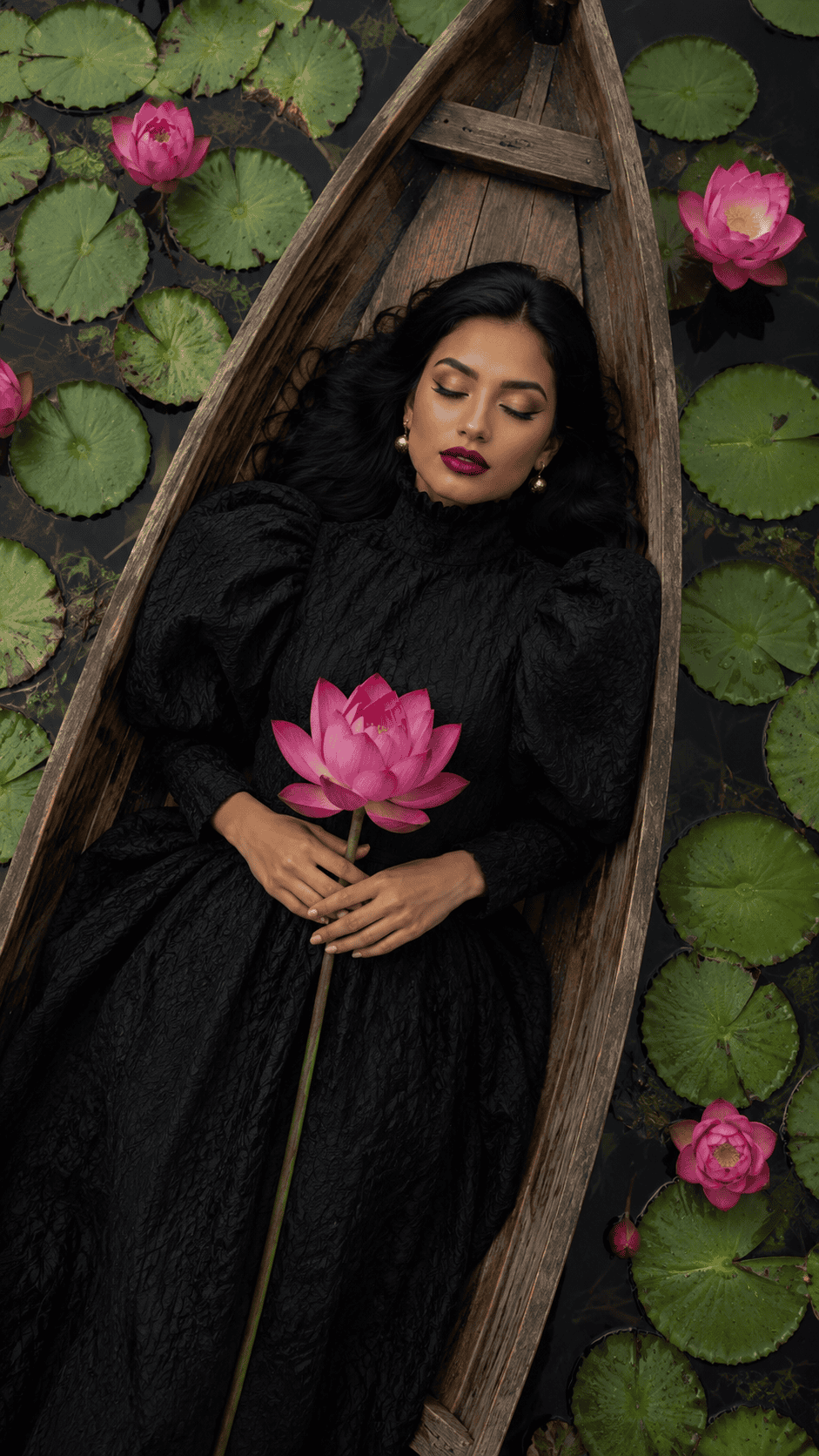 Woman in black dress holding a pink lotus in a wooden boat surrounded by water lilies.