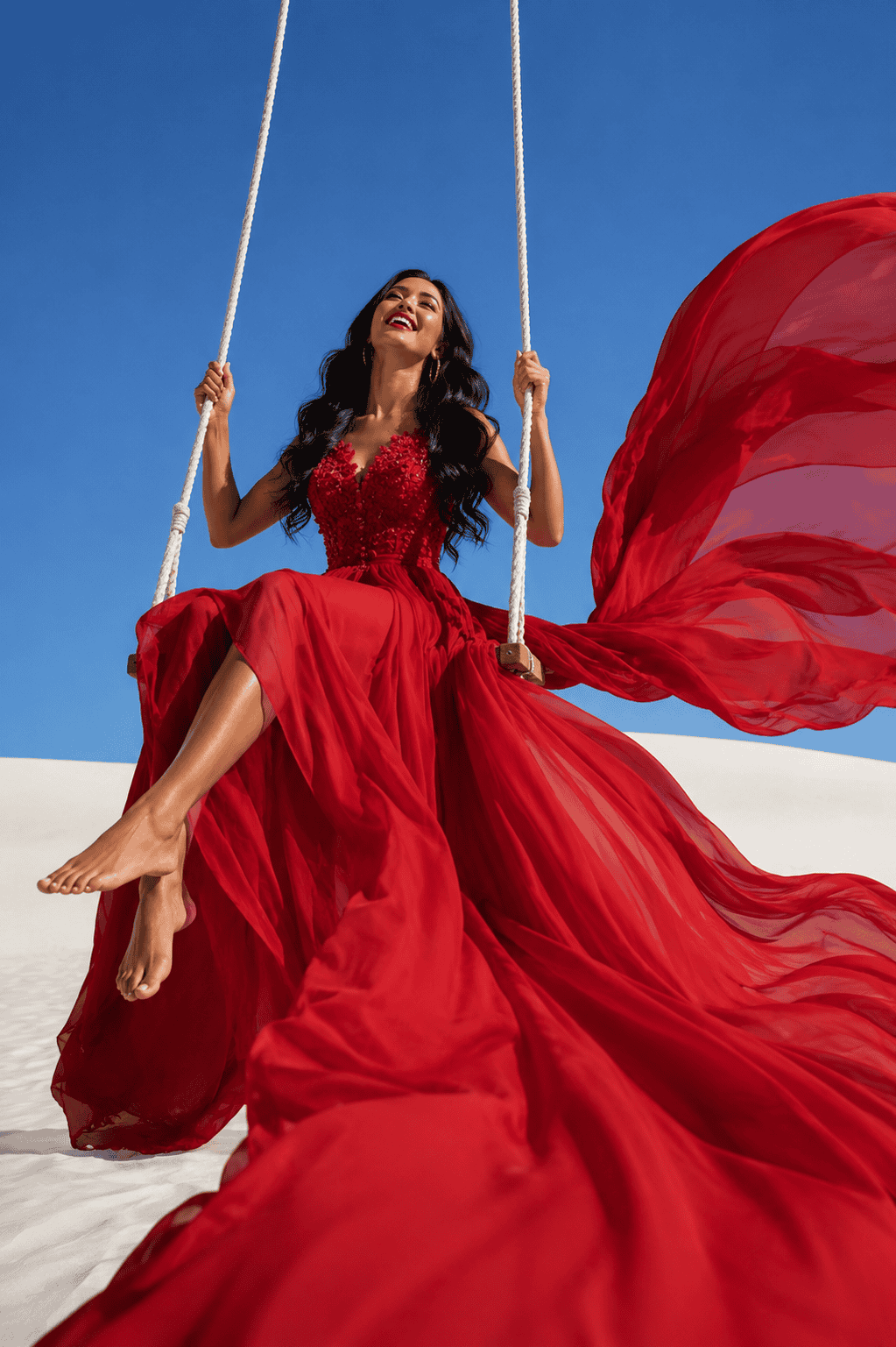 Woman in a flowing crimson gown sitting on a rope swing over white sand dunes, laughing joyfully under a clear blue sky with dramatic red fabric billowing in the wind.